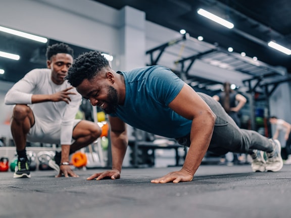 A man does push-ups in a gym as another observes, with medbridge digital care solutions implied for fitness support.