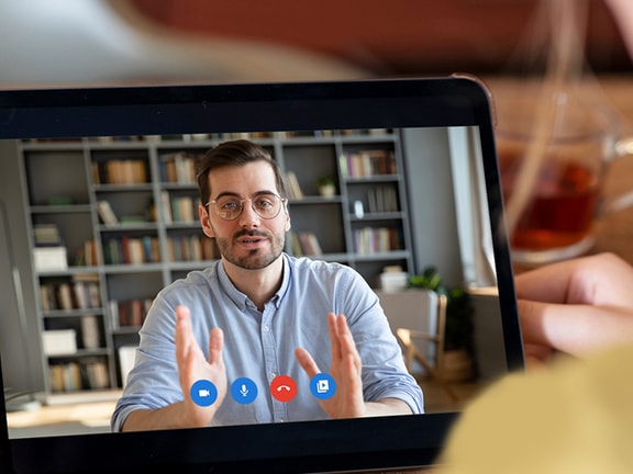 A video call on a tablet shows a man speaking, with bookshelves in the background, beside a tea cup and phone on the table.