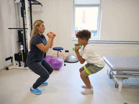 A physical therapist and a boy perform squats with dumbbells while using Medbridge patient care software.
