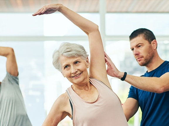 A Medbridge fitness instructor assists a senior woman with arm stretches, as an elderly man stretches nearby.