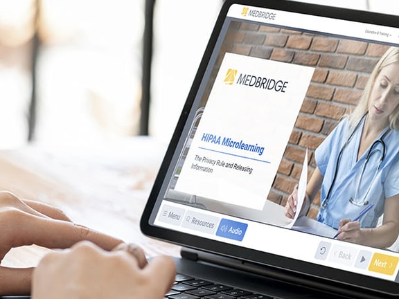 Laptop showing a healthcare-themed medbridge website with a doctor, beside a coffee cup and plant.