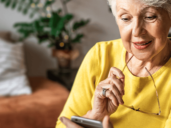 An older woman in a yellow sweater, holding glasses, smiles at her smartphone in a living room setting.
