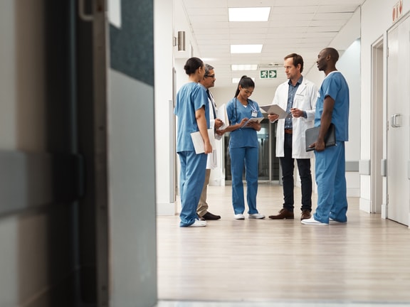 Group of clinicians in scrubs and a lab coat conferring over paperwork in a hospital corridor