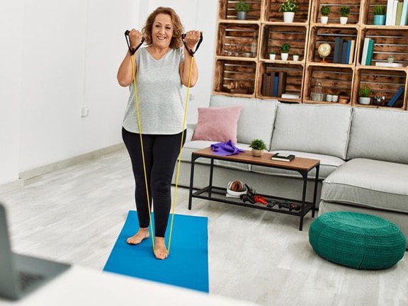 A woman exercises on a blue yoga mat in a bright living room with a laptop and furniture in the background.