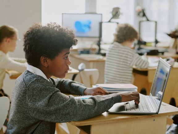 A student with curly hair types on a laptop at a classroom desk while classmates work nearby in the background.