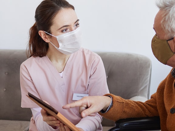 A nurse in pink and a mask holds a tablet, talking to an elderly masked man on a sofa at home.