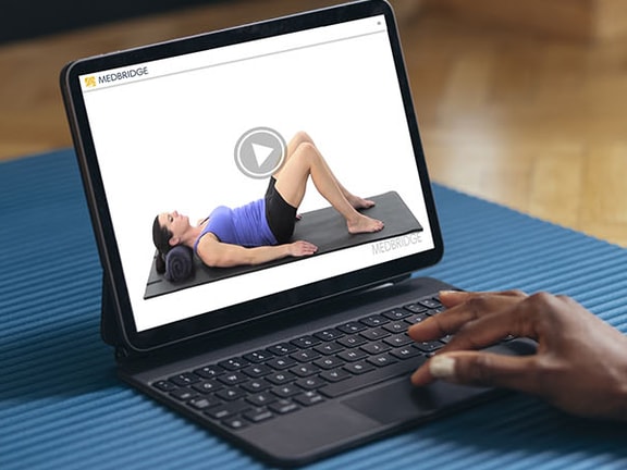 Person using a tablet to follow along with a home exercise program video on a yoga mat, illustrating HEP therapy.