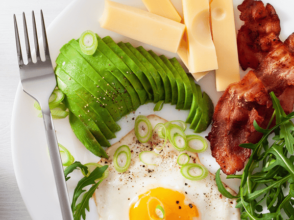 A breakfast plate with avocado, egg, bacon, arugula, cheese, plus an avocado half and juice nearby.
