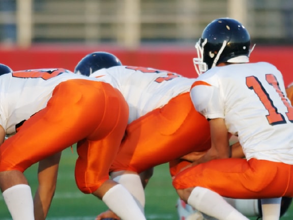 Football players in white and orange uniforms are lined up at the start of a game on Medbridge field.
