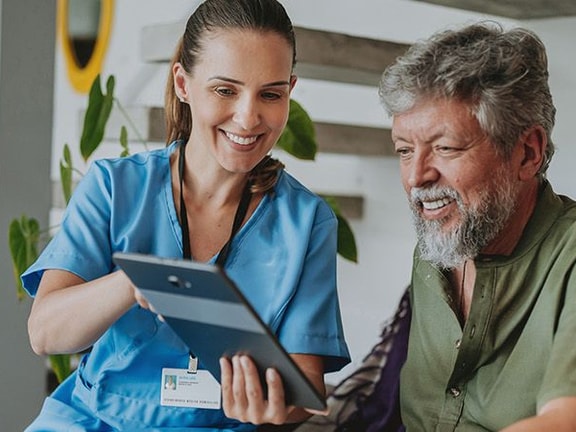 A healthcare worker and an older man smile while looking at a tablet in a bright room.