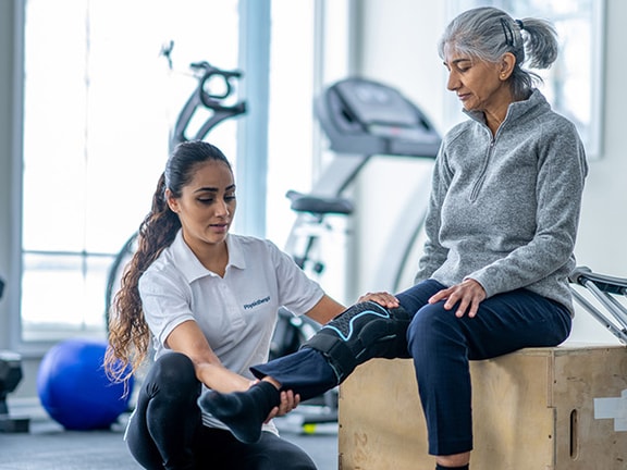 A Medbridge therapist helps an elderly woman with leg exercises in a gym.