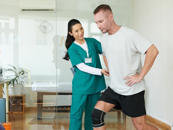 A physical therapist helps a man in a knee brace with balance exercises in a Medbridge therapy room.