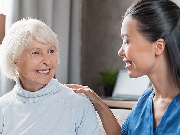 An elderly woman with white hair smiles at a younger woman in blue scrubs who is touching her shoulder.