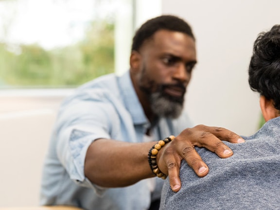 A bearded man offers comfort to someone beside a large window and beige couch in the medbridge room.