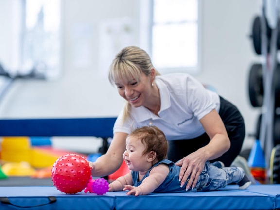 A woman uses medbridge digital healthcare to help a baby on a mat reach for red and purple toys in a therapy session.