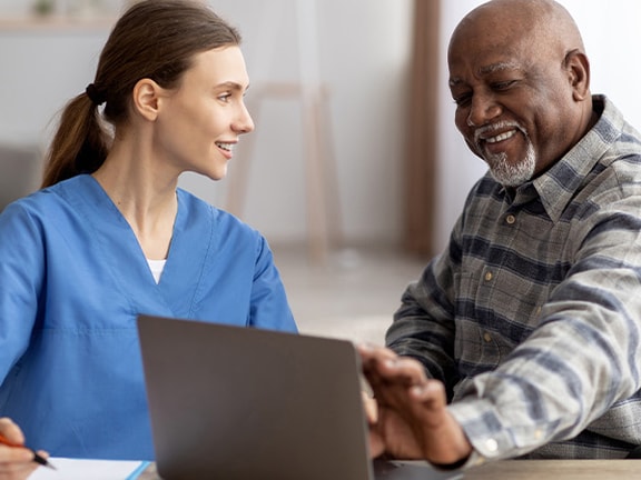 A Medbridge nurse in blue scrubs discusses a laptop screen with a man in a wheelchair.