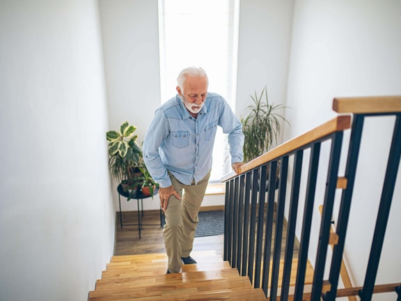Older adult using stair railing at home while managing knee pain, illustrating fall risk scenarios addressed with the ABC Scale PDF.