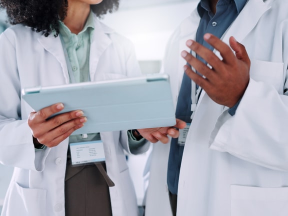 Two clinicians in lab coats review data on a tablet together while discussing a patient's care