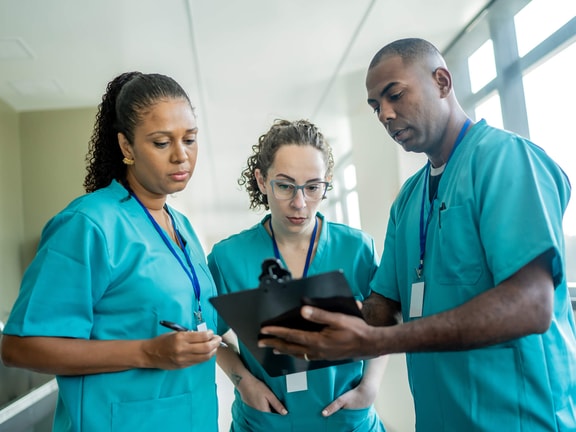 Three healthcare workers in scrubs review a clipboard, discussing training and medbridge patient care software in a hallway.