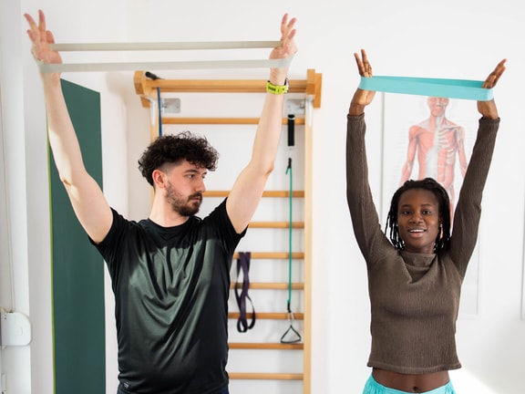 Two people exercise indoors with resistance bands, surrounded by equipment and a muscle anatomy poster in a medbridge training setting.