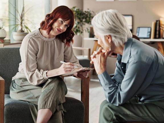 An occupational therapist holding a clipboard listens compassionately to an older patient during a behavioral health consultation.