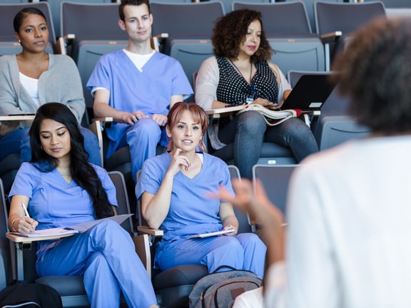 Students in scrubs sit in an auditorium listening to a lecture on new digital healthcare and patient care software.