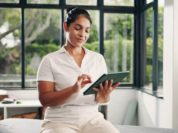 Clinician in a bright, plant-filled clinic sits on a treatment table, tapping a tablet to review patient outcomes