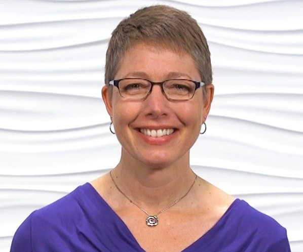 A woman with short hair and glasses, wearing a purple top, smiles in front of a white textured background, representing medbridge.