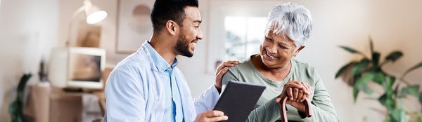 A doctor with a tablet interacts warmly with an elderly patient in a medbridge-style room.