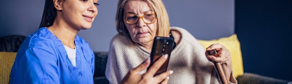 A caregiver in blue scrubs shows a smartphone to an elderly woman sitting on a couch, highlighting their use of medbridge for digital education.