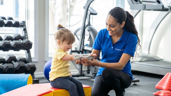 A woman and a young girl in a gym collaborate on an object; exercise equipment visible, medbridge context.