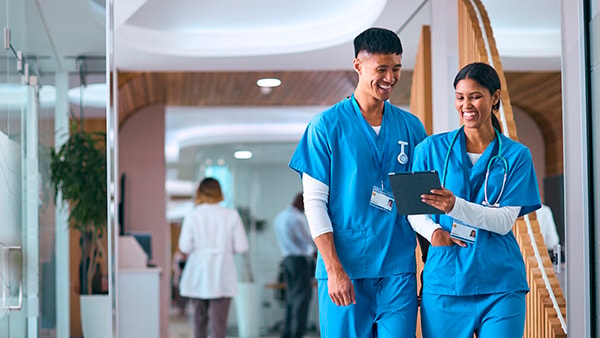 Two healthcare workers in blue scrubs smile at a tablet while walking through a hospital.