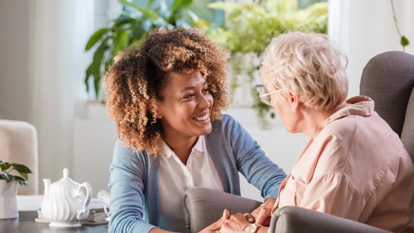 A woman with curly hair smiles at an older woman as they hold hands in a "warmly" lit room.