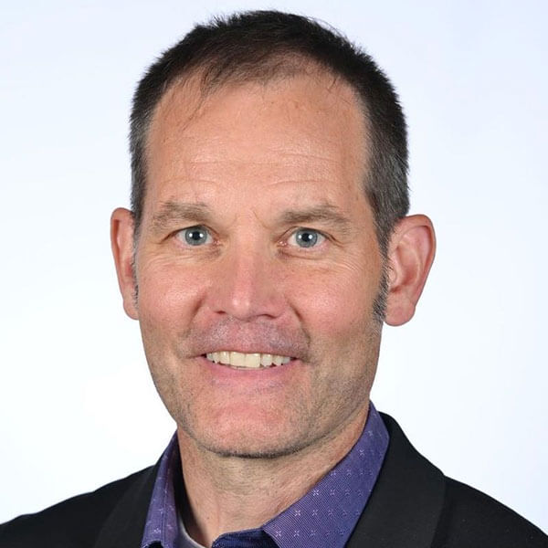 A person wearing a dark blazer and purple shirt is smiling at the camera.