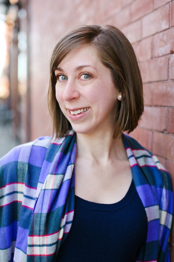 Person with short brown hair and a plaid shawl smiles in front of a brick wall.
