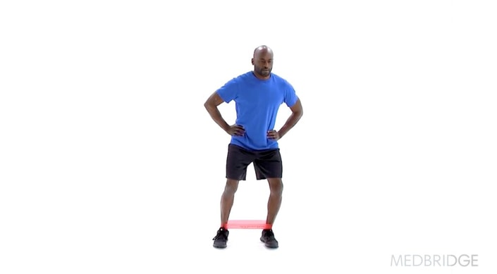 A person in a blue shirt and black shorts uses a red resistance band during exercise, integrating medbridge into their fitness routine.