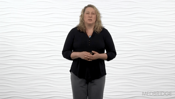 A person with wavy hair in a black top and gray pants stands against a white background.