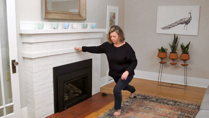 Middle-aged woman doing lunges with support from a white fireplace mantle, in a living room setting with plants and wall art, for medbridge.