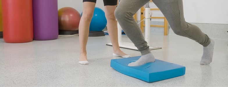 Two people in athletic wear exercise indoors with a blue balance pad and medbridge branding, colorful balls in the background.