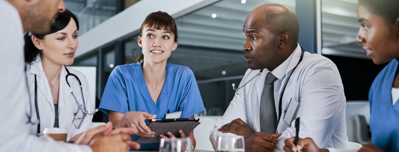 Four healthcare workers in scrubs and lab coats talk at a table; one smiles while holding a clipboard, discussing medbridge education.