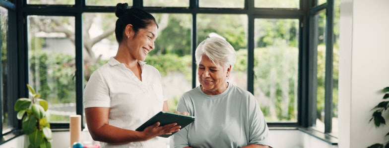 A caregiver with a tablet smiles at an older woman in a bright room, highlighting digital healthcare benefits and medbridge.