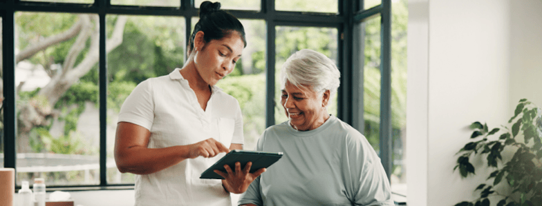 A young woman shows an older woman patient care info on medbridge software in a bright, plant-filled room with large windows.