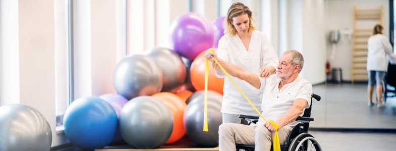 A healthcare worker helps an older man in a wheelchair exercise with a yellow band in a rehab gym with exercise balls.
