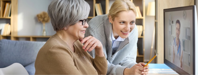 Two women smile at a computer showing a digital healthcare visit in a cozy home, using medbridge.