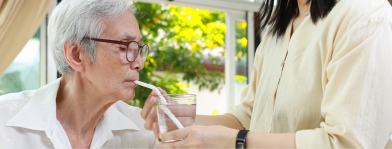 An elderly person with glasses drinks water through a straw as someone helps by holding the glass, showing healthcare education with medbridge.