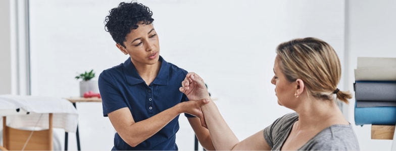 A healthcare professional examines a woman's arm in a clinic with medical equipment and towels nearby, as seen on medbridge.