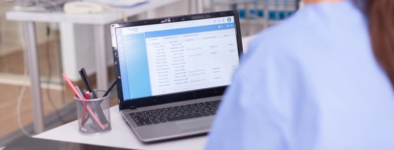 A person in scrubs sits at a desk viewing medical records on a laptop, with a pen holder nearby.