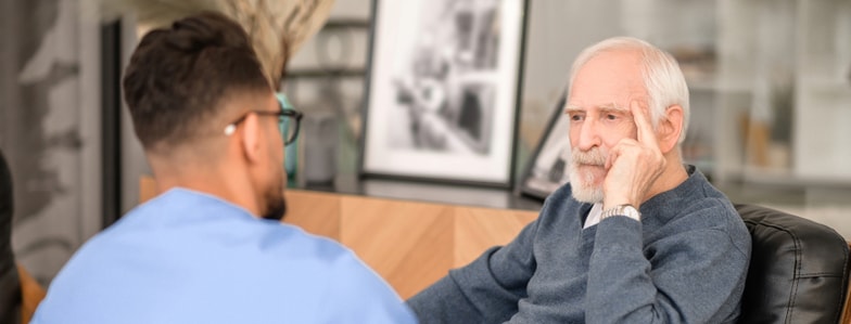 A senior man listens to a healthcare worker in blue scrubs indoors.