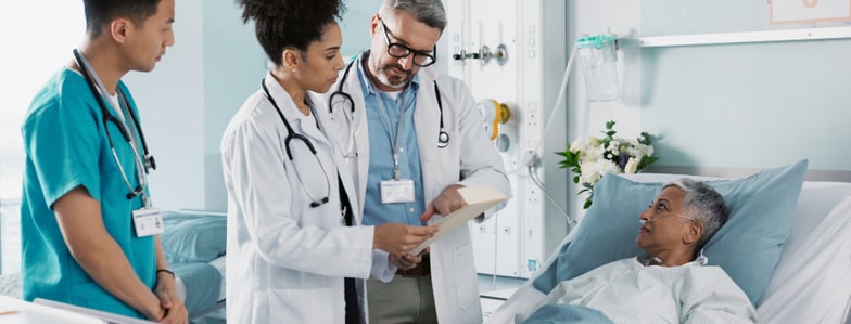 Three medical professionals use medbridge at a hospital bedside while an elderly patient watches.