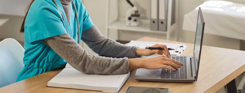 A person in medical scrubs works on a laptop at a desk with office items and a microscope, reflecting medbridge's digital healthcare setting.